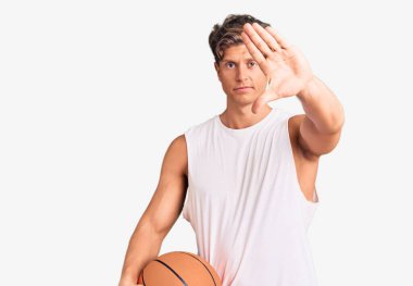 Genç yakışıklı adam basketbol topunu açık elle tutuyor dur işareti yapıyor ciddi ve kendinden emin bir ifadeyle, savunma hareketi yapıyor. 