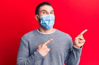 Tıbbi maske takan genç yakışıklı adam gülümsüyor ve kameraya bakıyor iki eliyle ve parmaklarıyla kenarı işaret ediyor.. 