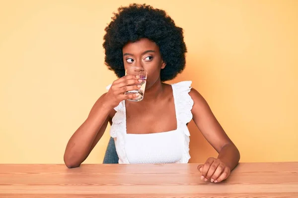 Young african american woman with afro hair drinking a glass of fresh water sitting on the table at home