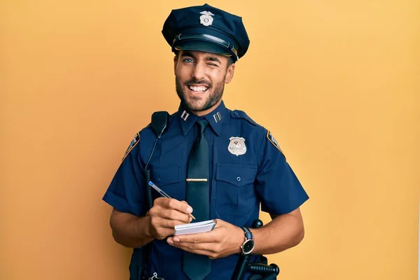Handsome hispanic man wearing police uniform writing traffic fine winking looking at the camera with sexy expression, cheerful and happy face.