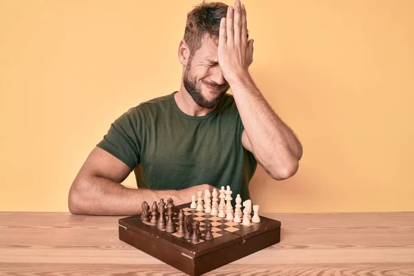 Young caucasian man playing chess sitting on the table surprised with hand on head for mistake, remember error. forgot, bad memory concept.
