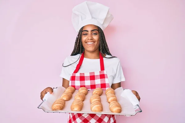 Young african american woman with braids wearing baker uniform holding homemade bread smiling and laughing hard out loud because funny crazy joke.