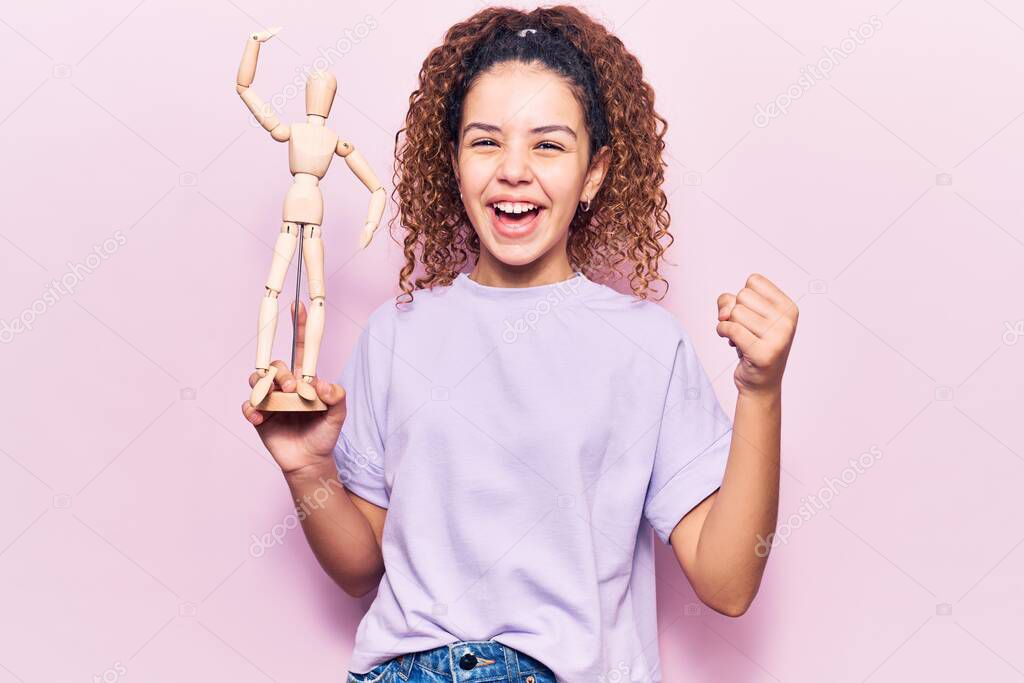 Beautiful kid girl with curly hair holding small wooden manikin screaming proud, celebrating victory and success very excited with raised arms 