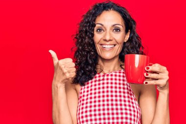 Orta yaşlı güzel bir kadın bir kupa kahve içiyor mutlu ve pozitif gülümsüyor, başparmağıyla mükemmel ve onay işareti yapıyor. 