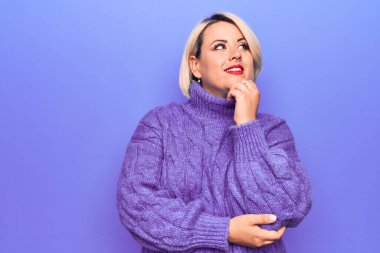 Güzel sarışın ve cüsseli bir kadın mor arka planda balıkçı yaka kazak giyiyor ve elleri çenesinde soru sormayı düşünüyor. Düşünceli bir ifade. Gülen ve düşünceli bir yüz. Şüphe kavramı.