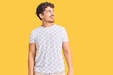 Kıvırcık saçlı, sıradan kıyafetler giyen, yüzünde gülümseme, doğal bir ifade olan genç yakışıklı bir adam. Kendinden emin gülen. 