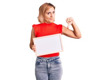 Kızgın yüzlü, boş beyaz karatahta tutan genç sarışın kadın, olumsuz işaret baş parmakları aşağıda, reddedilme konsepti 