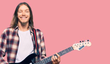 Uzun saçlı yakışıklı beyaz adam elektro gitar çalıyor. Pozitif görünüyor ve mutlu görünüyor. Kendine güvenen bir gülümsemeyle dişlerini gösteriyor. 