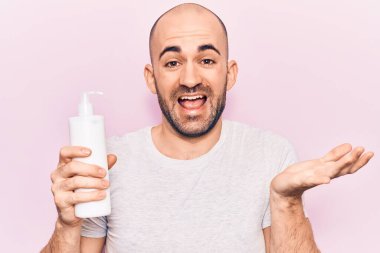 Genç yakışıklı kel adam elinde krem losyon şişesiyle başarıyı kutluyor mutlu bir gülümsemeyle ve kazanan yüz ifadesiyle elini kaldırarak. 