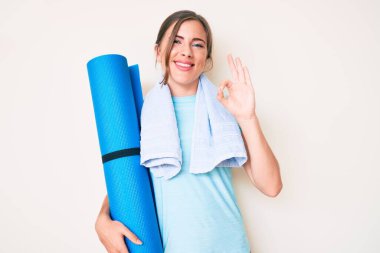 Güzel beyaz kadın yoga minderi tutuyor. Parmaklarıyla işaret veriyor. Arkadaşça gülümseyerek mükemmel bir sembol oluşturuyor. 