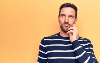 Sarı arka plan üzerinde duran sıradan çizgili süveter giyen genç ve yakışıklı bir adam parmak çenesinde ve yukarı bakarak şüphe üzerine yoğunlaşmıştı.