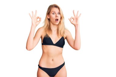 Bikini giyen güzel sarışın kadın şaşırmış ve şok olmuş görünüyor. Tamam sembolünü parmaklarıyla yapıyor. çılgın ifade 
