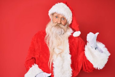 Gri saçlı, uzun sakallı, geleneksel Noel Baba kostümü giyen mutlu yüzüyle gülümseyen ve baş parmağıyla yüzünü gösteren yaşlı bir adam.. 