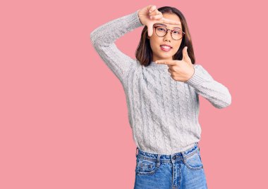 Genç, güzel Çinli bir kız günlük kıyafetler giyip gülümsüyor. Elleriyle ve parmaklarıyla gülen yüzüyle çerçeve yapıyor. yaratıcılık ve fotoğrafçılık kavramı. 