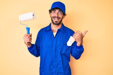 Kıvırcık saçlı, ayı kılıklı, inşaatçı tulumu giyen, elinde boya silindiri tutan, baş parmağını yana doğru uzatan ve ağzı açık gülümseyen yakışıklı bir genç. 