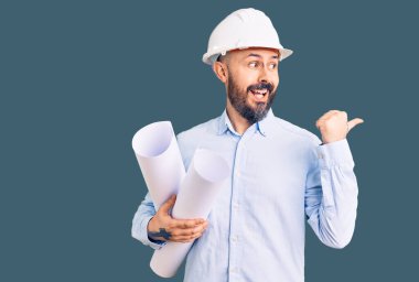 Elinde kağıt planlar tutan, kasketli genç yakışıklı bir adam başparmağıyla yan tarafa bakıyor, ağzı açık gülümsüyor. 