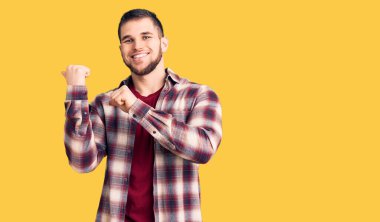 Gündelik tişört giyen genç bir adam arka tarafı işaret ediyor. El ve baş parmakları havada, kendinden emin gülümsüyor. 