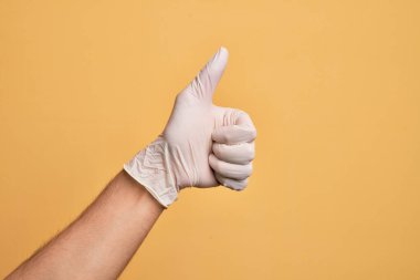 Sarı arka plan üzerinde tıbbi eldiveni olan beyaz bir gencin eli başarılı bir onay hareketi yapıyor. Başparmak işareti, onaylama ve olumlu bir sembol.