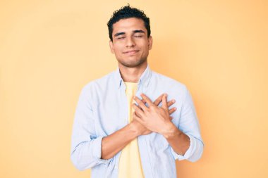 Sarı arka planda duran yakışıklı İspanyol adam elleri göğsünde, gözleri kapalı gülümsüyor ve yüzünde minnet ifadesi var. sağlık kavramı. 