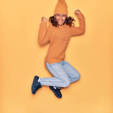 Yün şapka ve süveter giyen, mutlu gülümseyen genç, güzel Latin bir kadın. Yüzünde tebessümle zıplıyor, sarı arka planda yumruklar havada kutluyor.