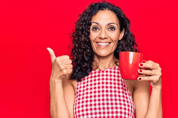 Orta yaşlı güzel bir kadın bir kupa kahve içiyor mutlu ve pozitif gülümsüyor, başparmağıyla mükemmel ve onay işareti yapıyor. 