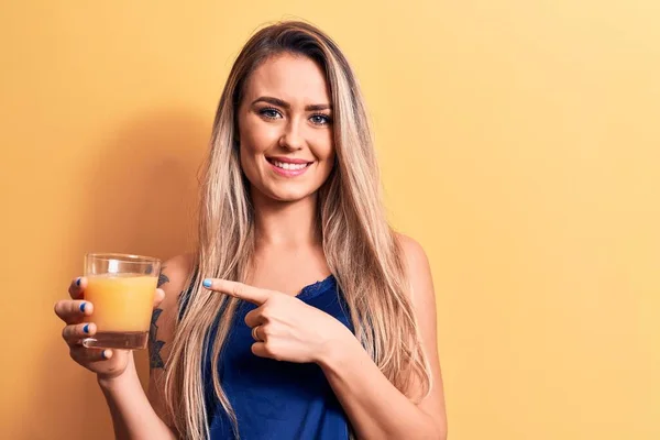 年轻美丽的金发女子在黄色的背景下喝着一杯健康的橙汁 高兴地微笑着 手指手画脚 — 图库照片