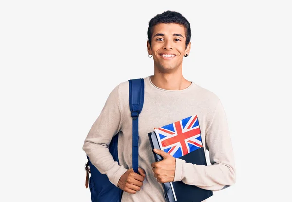 若いアフリカ系アメリカ人の男を着て学生のバックパックを保持バインダーと英国のフラグを見て正と幸せな立ちと笑顔で自信を持って笑顔を示す歯 — ストック写真