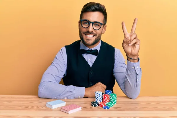 英俊而惊慌失措的男人坐在桌子上 手里拿着扑克牌和卡片 开心地微笑着 在镜头前眨眼示意胜利 第二点 — 图库照片