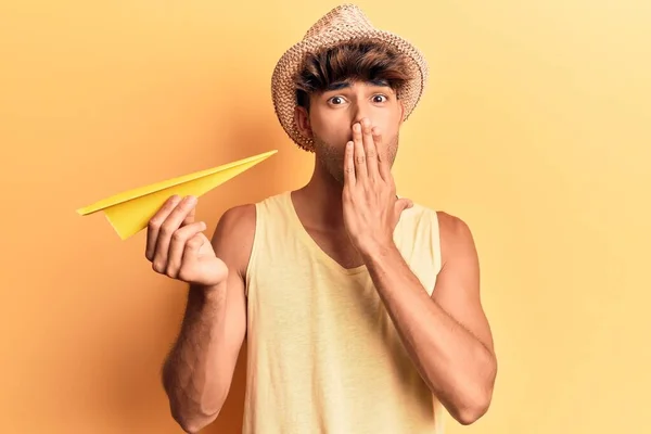 夏の帽子をかぶった若いヒスパニック系の男性は 手で口をカバー紙飛行機を保持し ショックを受け 間違いを恐れています 驚きの表情 — ストック写真