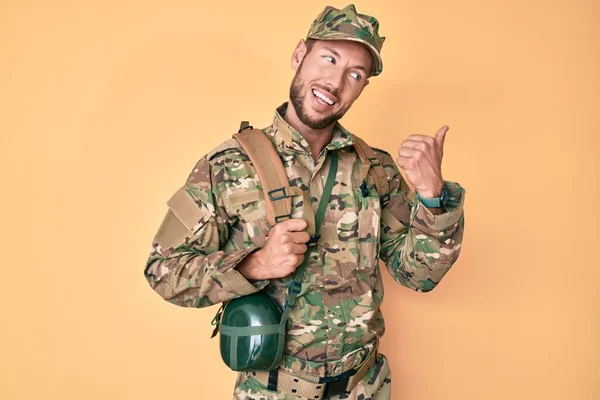 Young caucasian man wearing camouflage army uniform and canteen pointing thumb up to the side smiling happy with open mouth