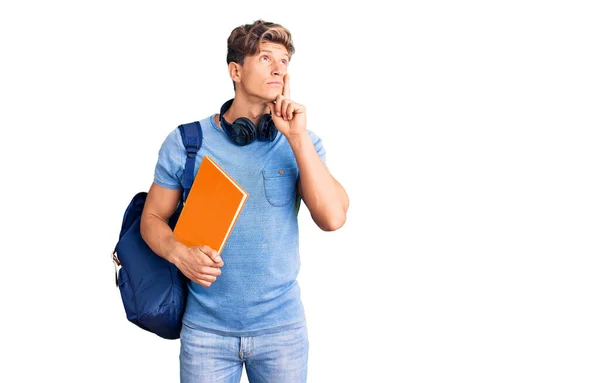 年轻英俊的男子背着学生背包 头戴耳机 手里拿着书本 严肃地看待问题 双手托着下巴 沉思着困惑的想法 — 图库照片