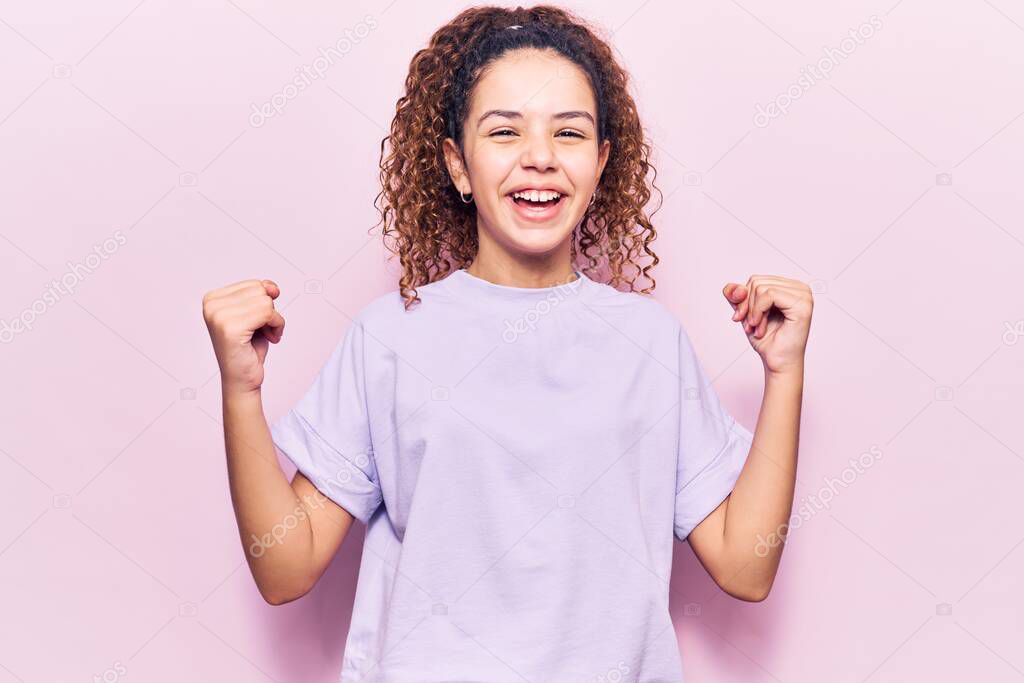 Beautiful kid girl with curly hair wearing casual clothes screaming proud, celebrating victory and success very excited with raised arms 
