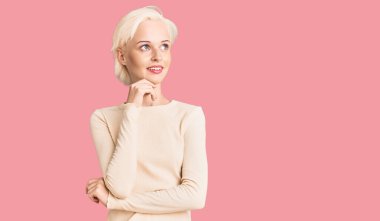 Elleri çenesinde gündelik giysiler giymiş soru soran genç sarışın kadın, düşünceli bir ifade. Gülümseyen ve düşünceli bir yüz. şüphe kavramı. 