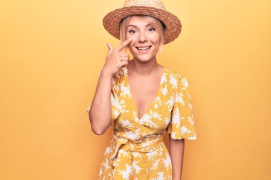 Yaz şapkası takıp sarı arka planda giyinen güzel sarışın kadın. El parmağı ve burunla işaret ediyor. Neşeli gülümsüyor. Güzellik konsepti