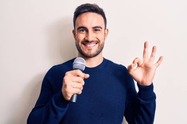 Beyaz arka planda mikrofon kullanarak şarkı söyleyen sakallı yakışıklı genç şarkıcı parmaklarıyla imza atıp gülümseyerek mükemmel bir sembol oluşturuyor.