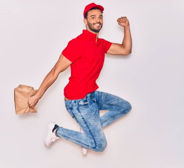 Mutlu gülümseyen üniformalı yakışıklı İspanyol teslimatçı. Yüzünde tebessümle, elinde torbayla, izole edilmiş beyaz arka planda paket yiyeceklerle zıplıyorsun..