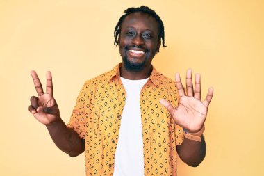 Saçları örgülü genç Afro-Amerikan bir adam 7 numaralı parmaklarını gösterip gösterirken kendinden emin ve mutlu bir şekilde gülümsüyor.. 