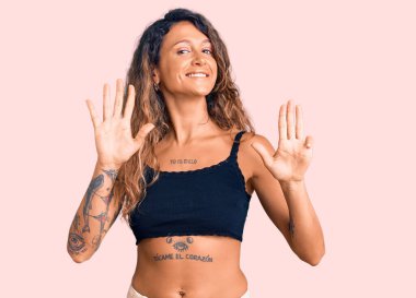 Dövmeli genç İspanyol kadın. Günlük giysiler giyiyor. Dokuz numaralı parmaklarıyla işaret ediyor ve gülümsüyor.. 