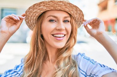 Yaz şapkası takmış, mutlu bir şekilde gülümseyen güzel sarışın bir kadın. Şehrin caddesinde yüzünde gülümsemeyle dikilirken.