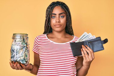 Tasarruf kavanozu ve cüzdanı olan, yüzünde ciddi bir ifadeyle rahatlamış genç Afrikalı Amerikalı kadın. Basit ve doğal bir şekilde kameraya bakıyorsun.. 