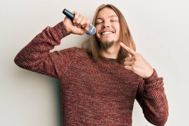 Uzun saçlı yakışıklı beyaz adam mikrofon kullanarak şarkı söylüyor mutlu bir şekilde eli ve parmağı işaret ediyor. 