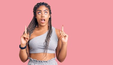 Spor kıyafetleri giymiş Afro-Amerikalı genç bir kadın şaşırmış ve şaşırmış bir şekilde yukarı bakıyor ve parmaklarını gösterip kollarını kaldırıyor.. 