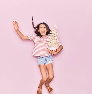 Tatlı İspanyol kız ağzı açık elinde patlamış mısırla izole edilmiş pembe arka plandan atlarken şaşırdı.