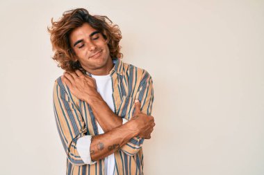 Gündelik kıyafetler giyen genç İspanyol adam kendine sarılıyor mutlu ve pozitif, kendinden emin gülümsüyor. Sevgi ve özgüven. 