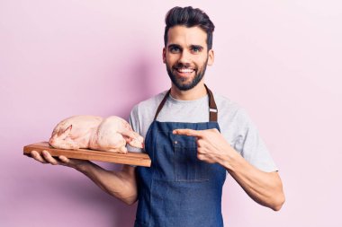 Sakallı, önlüklü, çiğ tavuklu, gülümseyen eli ve parmağı olan genç yakışıklı bir adam. 