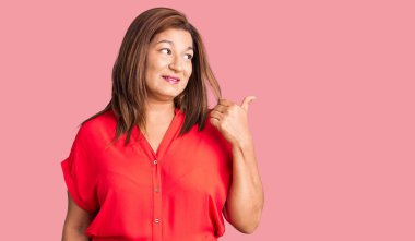 Orta yaşlı, sıradan kıyafetler giyen, mutlu yüzlü gülümseyen ve baş parmağıyla yüzünü gösteren Latin bir kadın.. 