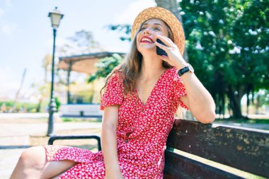 Genç sarışın kadın tatilde. Parktaki bankta oturmuş akıllı telefondan konuşuyor.