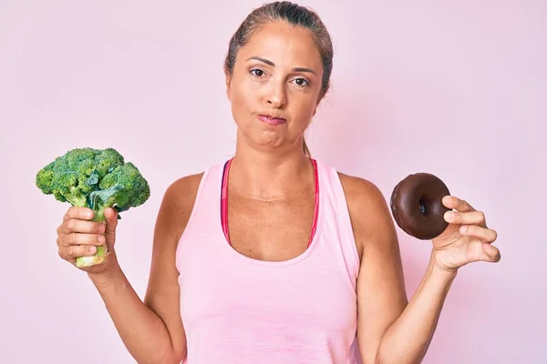 中年时 他惊慌失措的女人抱着花椰菜和巧克力甜甜圈 怀疑和紧张 皱着眉头因为问题而烦恼 消极的人 — 图库照片