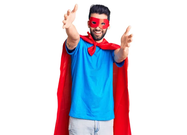 Young handsome man with beard wearing super hero costume looking at the camera smiling with open arms for hug. cheerful expression embracing happiness.