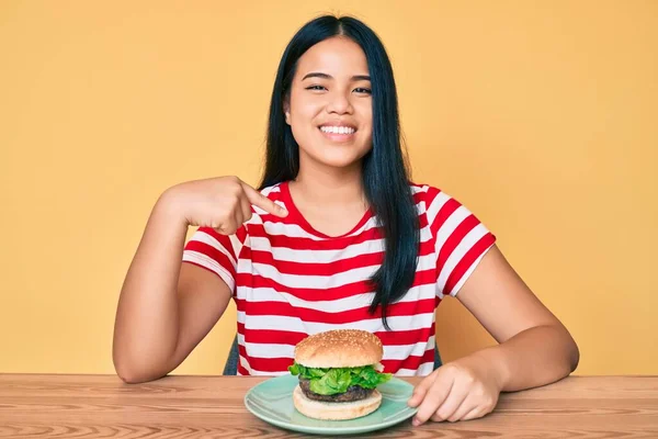 Fiatal Ázsiai Lány Eszik Egy Ízletes Klasszikus Burger Ujjal Mutogatva — Stock Fotó